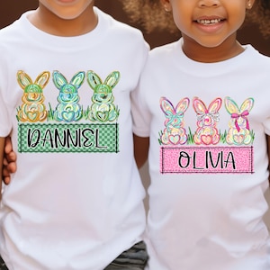 May include: White t-shirts with colorful Easter bunny graphics. One shirt features the name "DANIEL" on a green checkered background, while the other says "OLIVIA" on a pink leopard print background. The bunnies have heart-shaped bodies.