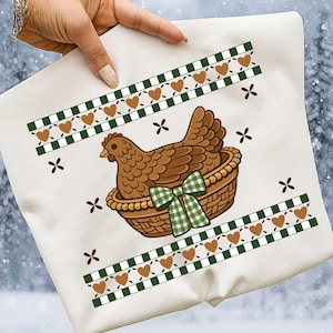 Może przedstawiać: Biała kwadratowa poduszka z brązową kurą w wiklinowym koszu, ozdobiona zieloną kokardą w kratkę. Projekt zawiera brązowe serca, zielono-białe obramowania w kratkę i małe brązowe kształty krzyży.