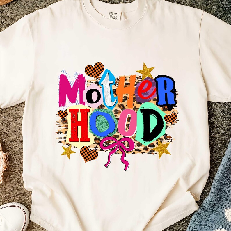 May include: Cream-colored t-shirt with the word "MOTHERHOOD" in colorful, playful lettering. The design includes hearts, stars, and a pink bow, set against a leopard print background. A fun, graphic tee.