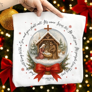 May include: White Christmas-themed pillow featuring a snow globe design with the Nativity scene inside. The snow globe is set on a red bow. Text includes "And you shall call His name Jesus, for He will save His people from their sins. Matthew 1:21."