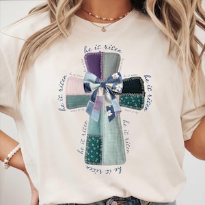 Könnte beinhalten: Cremefarbenes T-Shirt mit einem Patchwork-Kreuz-Design und einer blauen Schleife. Das Kreuz besteht aus verschiedenen Stoffmustern in Lila-, Grün- und Blautönen. Die Worte "he is risen" sind in einer Schreibschrift um das Kreuz geschrieben.