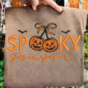 May include: Brown tote bag with orange embroidered text and graphics. The text reads "Spooky Season" with two jack-o'-lanterns and two bats. The bag is held up by a hand.