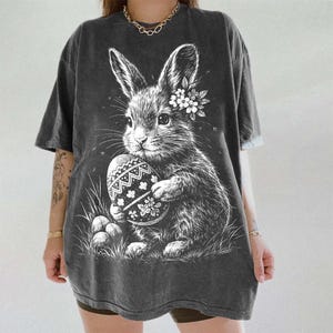 May include: Dark gray t-shirt featuring a detailed illustration of an Easter bunny holding a decorated egg. The bunny has a floral crown and is surrounded by grass and additional eggs. The design is in white.