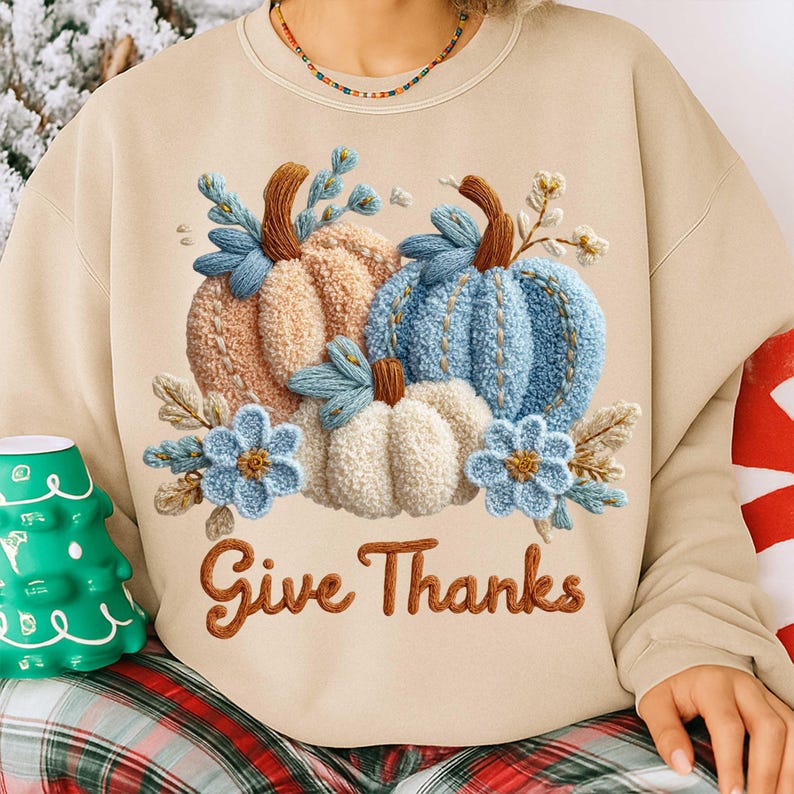 May include: Beige sweatshirt featuring embroidered pumpkins in shades of peach, blue, and white, with blue floral accents. The phrase "Give Thanks" is embroidered in brown script, perfect for fall and Thanksgiving.