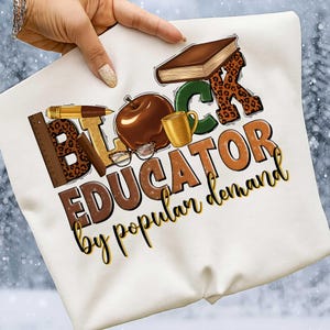 May include: White sweatshirt featuring the text "BLACK EDUCATOR by popular demand" in a colorful design with a ruler, pen, apple, coffee cup, book, and glasses. The letters "B" and "R" have a leopard print pattern.