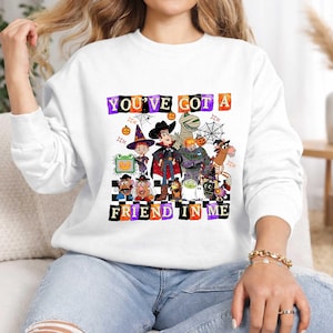 May include: A white long-sleeve sweatshirt featuring a graphic print of various animated characters dressed in Halloween costumes. The text "YOU'VE GOT A FRIEND IN ME" is displayed in colorful, blocky letters above and below the characters.