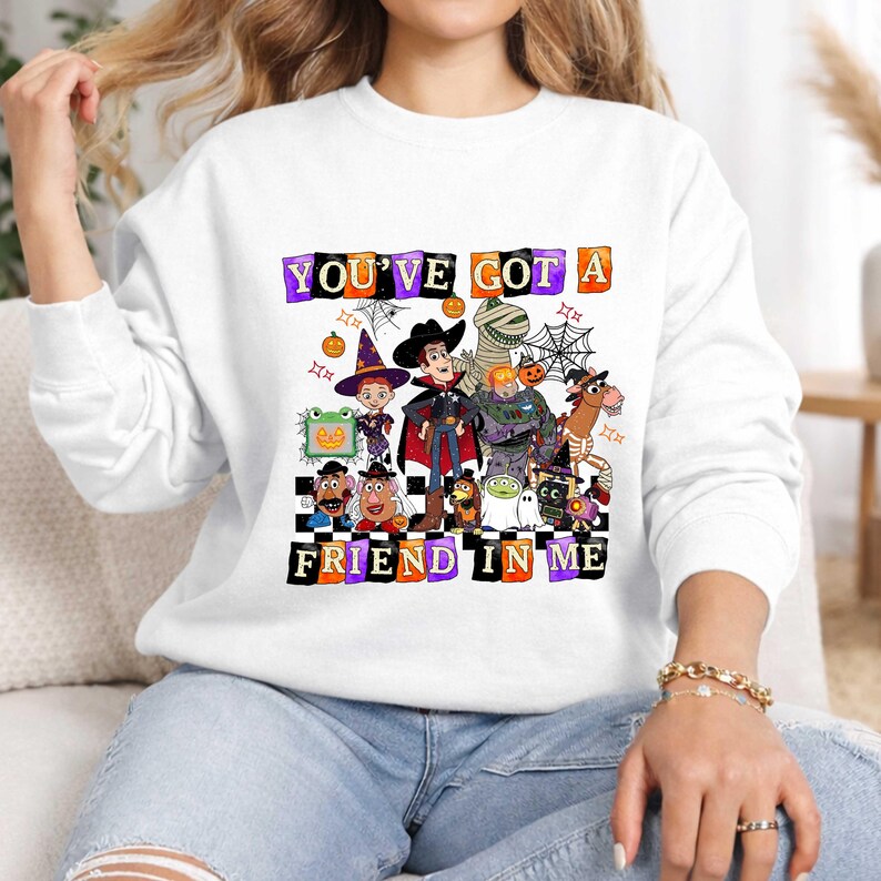May include: A white long-sleeve sweatshirt featuring a graphic print of various animated characters dressed in Halloween costumes. The text "YOU'VE GOT A FRIEND IN ME" is displayed in colorful, blocky letters above and below the characters.