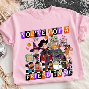 May include: A light pink t-shirt featuring a graphic print of various animated characters dressed in Halloween costumes. The text "YOU'VE GOT A FRIEND IN ME" is printed in colorful, blocky letters above and below the character illustration.