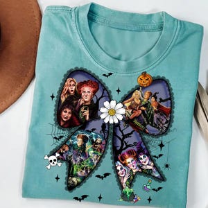 Op de afbeelding: Lichtblauw t-shirt met een groot strikdesign met afbeeldingen van heksen en Halloween-elementen. De strik is versierd met een witte madelief, vleermuizen en een schedel. Het shirt is een comfortabele, casual stijl.