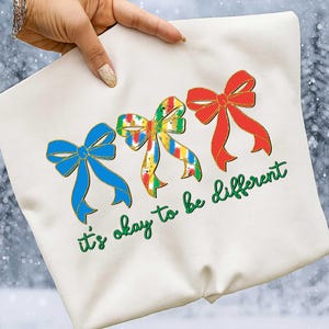 May include: Cream-colored sweatshirt featuring three decorative bows in blue, multicolored, and red with gold outlines. The phrase "it's okay to be different" is written in green script below the bows. The sweatshirt is held up by a person.