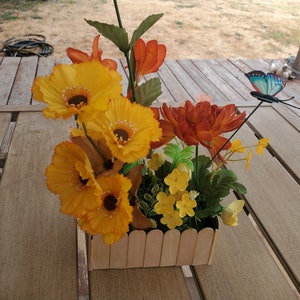 May include: A wooden planter box with a picket fence design, filled with artificial flowers. The flowers are yellow, orange, and red, and there is a blue butterfly on a wire stem.