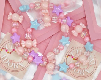 Puede incluir: Un kit de elaboración de joyas rosa y azul con cuentas con forma de caramelo, estrella y flor. El kit incluye una bolsa de plástico transparente con las cuentas y una tarjeta rosa y blanca con las palabras "Kits de Joyas" y "Charm Idol" impresas en ella.