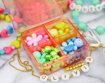 Puede incluir: Un recipiente de plástico transparente con seis compartimentos llenos de cuentas de colores. El recipiente está abierto y las cuentas son visibles. Un collar de cadena dorada con cuentas de letras blancas que deletrean "OLIVIA" está en primer plano.