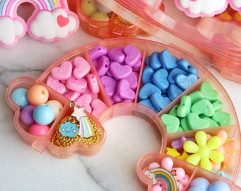 May include: A rainbow-shaped plastic container with multiple compartments filled with colorful beads, charms, and other craft supplies. The container is open and the compartments are visible. The beads are in various shapes, including hearts, stars, and flowers. The charms include a rainbow, a cloud, and a star.