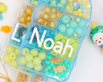 Puede incluir: Un recipiente de plástico azul con compartimentos llenos de cuentas y abalorios de colores. El recipiente tiene forma de helado de paleta y lleva el nombre "Noah" escrito.