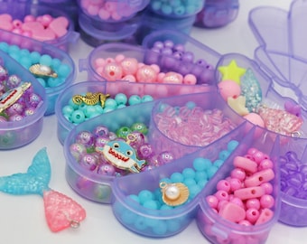 Puede incluir: Una colección de cuentas y abalorios morados, rosas y azules en recipientes de plástico transparente. Los recipientes están divididos en secciones e incluyen cuentas de varios tamaños y formas, junto con dijes de cola de sirena y conchas marinas.