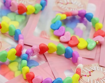 May include: A close-up of three rainbow heart-shaped beaded bracelets. The bracelets are made of colorful plastic beads and are arranged on a pink background. The bracelets are packaged in clear plastic bags with pink labels that say "Charm Addict Jewelry".