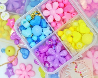 Puede incluir: Una caja de almacenamiento de plástico transparente con seis compartimentos llenos de cuentas de plástico de colores. Las cuentas tienen forma de estrellas, corazones, flores y círculos. La caja está etiquetada como "Clara's Jewelry Kids".