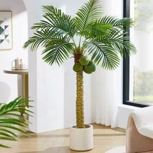 Può includere: Palma artificiale con foglie verdi e tronco marrone. L'albero ha un vaso bianco e diverse noci di cocco marroni appese ai rami.