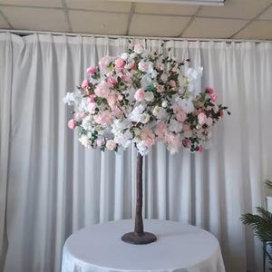 May include: A white and pink artificial flower tree centrepiece with a brown wooden base. The tree is decorated with white and pink flowers and is perfect for a wedding or other special event.