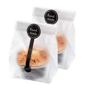 Pode incluir: Duas sacolas de papel brancas com bolinhas pretas, cada uma selada com um adesivo preto que diz "hand made". As sacolas contêm produtos de panificação. As sacolas são parcialmente transparentes, permitindo vislumbrar o conteúdo.