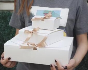 Könnte beinhalten: Drei weiße Geschenkboxen, die übereinander gestapelt sind, jede mit einem beigefarbenen Band gebunden. Die Boxen sind rechteckig und unterschiedlich groß, wobei die größte Box unten und die kleinste oben ist. Die Boxen werden von einer Person gehalten, die ein graues T-Shirt trägt.