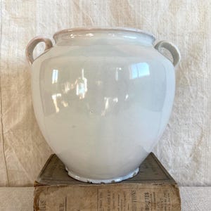 May include: A large, off-white ceramic vase with two curved handles. The vase has a rounded body and a slightly flared rim. It sits atop a stack of old books with faded text.