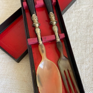 Peut inclure: Un ensemble de service vintage avec une cuillère et une fourchette, présenté dans une boîte noire avec un intérieur rouge. Les manches sont noirs, avec des détails en métal argenté orné. La cuillère est ambrée et la fourchette a trois dents.