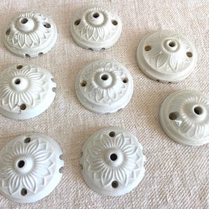 Puede incluir: Un conjunto de siete rosetones de techo circulares de cerámica color blanco roto. Cada pieza presenta un patrón floral en relieve que irradia desde un orificio central, con orificios adicionales situados alrededor del perímetro.