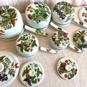 May include: A collection of white ceramic cookware with lids and handles, decorated with botanical illustrations of berries and leaves in red, green, and black. The set includes pots and pans of various sizes.