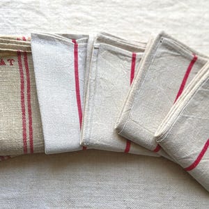May include: A stack of folded linen towels in various shades of beige and off-white. Each towel features a bold red stripe or stripes. Some towels have the embroidered letters "AT".