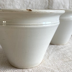 May include: Two white ceramic plant pots with a slightly flared shape. The pots have a smooth, glossy finish and a simple, classic design. The rim of the pot has a subtle decorative detail. Suitable for indoor or outdoor use, these pots are ideal for plants.