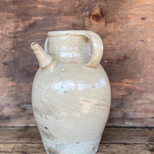 May include: A beige ceramic pitcher with a spout and handle. The jug has a rounded body and a light-colored, textured surface. The spout is small and angled, and the handle is curved. The pitcher is likely for decorative use.