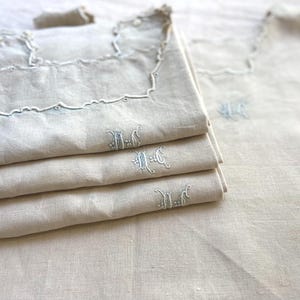 May include: A stack of three folded beige linen napkins with white embroidered scalloped edges and a white monogrammed letter M on the front. Beside the stack lies a single matching unfolded napkin featuring the same monogram.