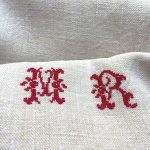 May include: Close-up of a beige linen fabric with two embroidered red initials, "MR". The letters are in a decorative, ornate style. The fabric has a subtle texture and a natural, neutral color.