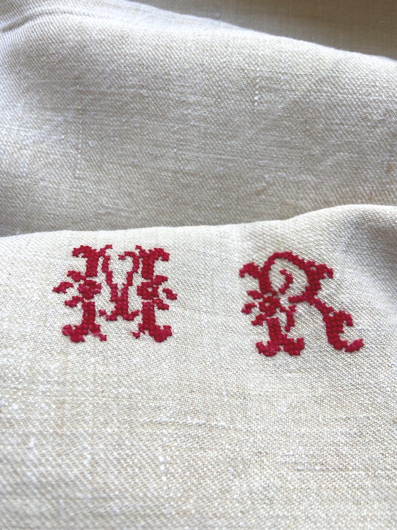May include: Close-up of a beige linen fabric with two embroidered red initials, "MR". The letters are in a decorative, ornate style. The fabric has a subtle texture and a natural, neutral color.