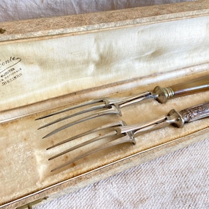 Peut inclure: Un ensemble de fourchettes de service en argent ancien, comprenant deux fourchettes avec des manches décoratifs, reposant dans un coffret doublé de velours crème. Le coffret a un fermoir doré et le texte "A. Conte, Bijoutier, Rue Nationals, Beziers" est imprimé à l'intérieur.