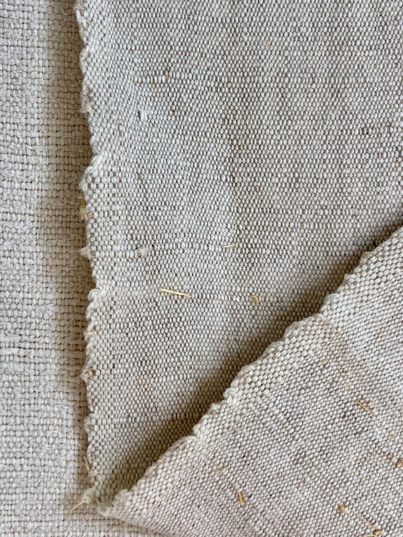 May include: Close-up of a piece of off-white woven fabric with a rough texture. The fabric is folded over itself, showing the raw edges.