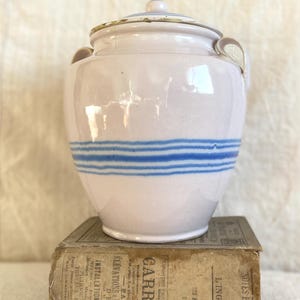 May include: A vintage ceramic jar with a lid, pale pink in colour, and three blue horizontal stripes. The jar has two small handles and sits on an old book with faded text. The lid is also pink.