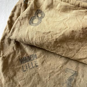 May include: Close-up of tan fabric bags with drawstring closures. One bag has a large black number 8 printed on it. Another bag has the text "WAREIN LILLE" printed in black. The bags appear to be vintage or antique.