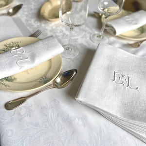 May include: A formal table setting with white linen napkins embroidered with the letters "EL". The napkins are placed on plates with a floral design, alongside silverware and wine glasses. The tablecloth has a subtle floral pattern.