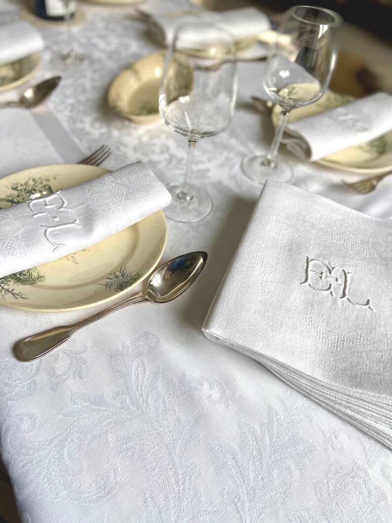 May include: A formal table setting with white linen napkins embroidered with the letters "EL". The napkins are placed on plates with a floral design, alongside silverware and wine glasses. The tablecloth has a subtle floral pattern.