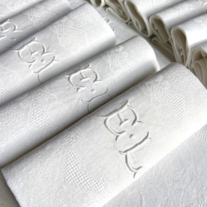 May include: A collection of white linen napkins, some rolled and others folded, with an embroidered monogram. The napkins have a subtle floral pattern and are arranged on a white surface, suggesting a formal dining setting.