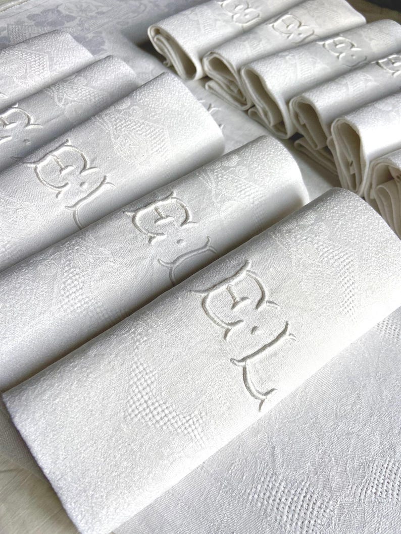 May include: A collection of white linen napkins, some rolled and others folded, with an embroidered monogram. The napkins have a subtle floral pattern and are arranged on a white surface, suggesting a formal dining setting.