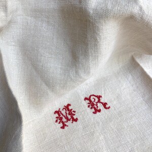 May include: Close-up of a cream-colored linen textile with red embroidered initials "MR". The fabric has a subtle texture and is slightly wrinkled. The embroidery is in a decorative, ornate style.