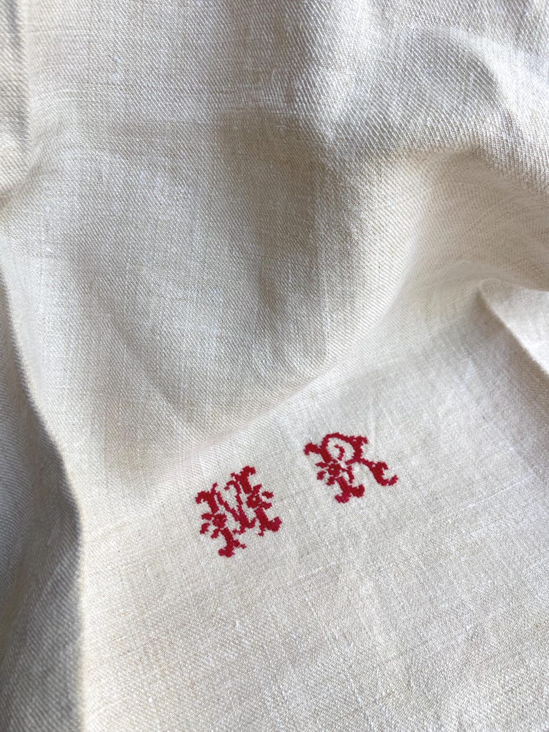 May include: Close-up of a cream-colored linen textile with red embroidered initials "MR". The fabric has a subtle texture and is slightly wrinkled. The embroidery is in a decorative, ornate style.