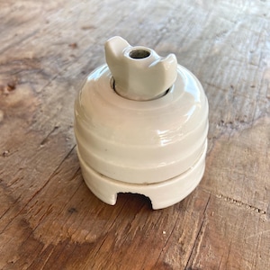 Op de afbeelding: Een vintage gebroken witte keramische draaischakelaar met een glanzende afwerking. De schakelaar heeft een centrale vleugelvormige knop op een ronde basis, met een kleine inkeping aan de onderkant van de behuizing voor de draadtoegang.