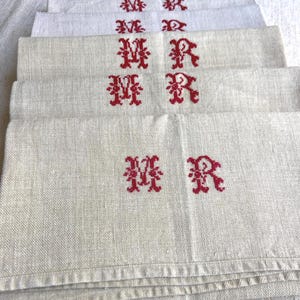 May include: A stack of off-white linen napkins, each embroidered with a red "MR" monogram. The napkins have a textured appearance and are neatly folded, showcasing the embroidered initials prominently. The overall aesthetic is vintage and classic.