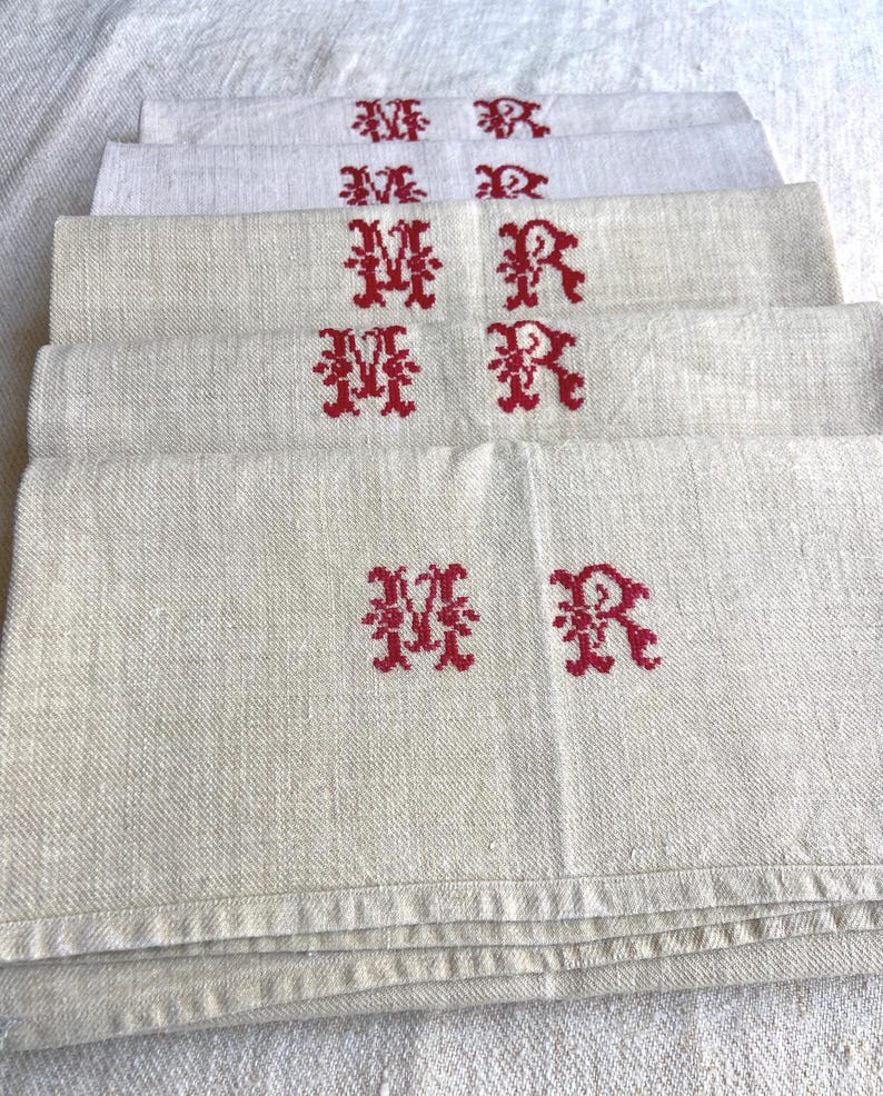 May include: A stack of off-white linen napkins, each embroidered with a red "MR" monogram. The napkins have a textured appearance and are neatly folded, showcasing the embroidered initials prominently. The overall aesthetic is vintage and classic.