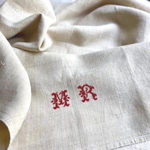 May include: Cream-colored linen cloth with red embroidered initials "MR". The fabric is folded, showing texture and slight variations in color. The embroidery is in a decorative, stylized font.